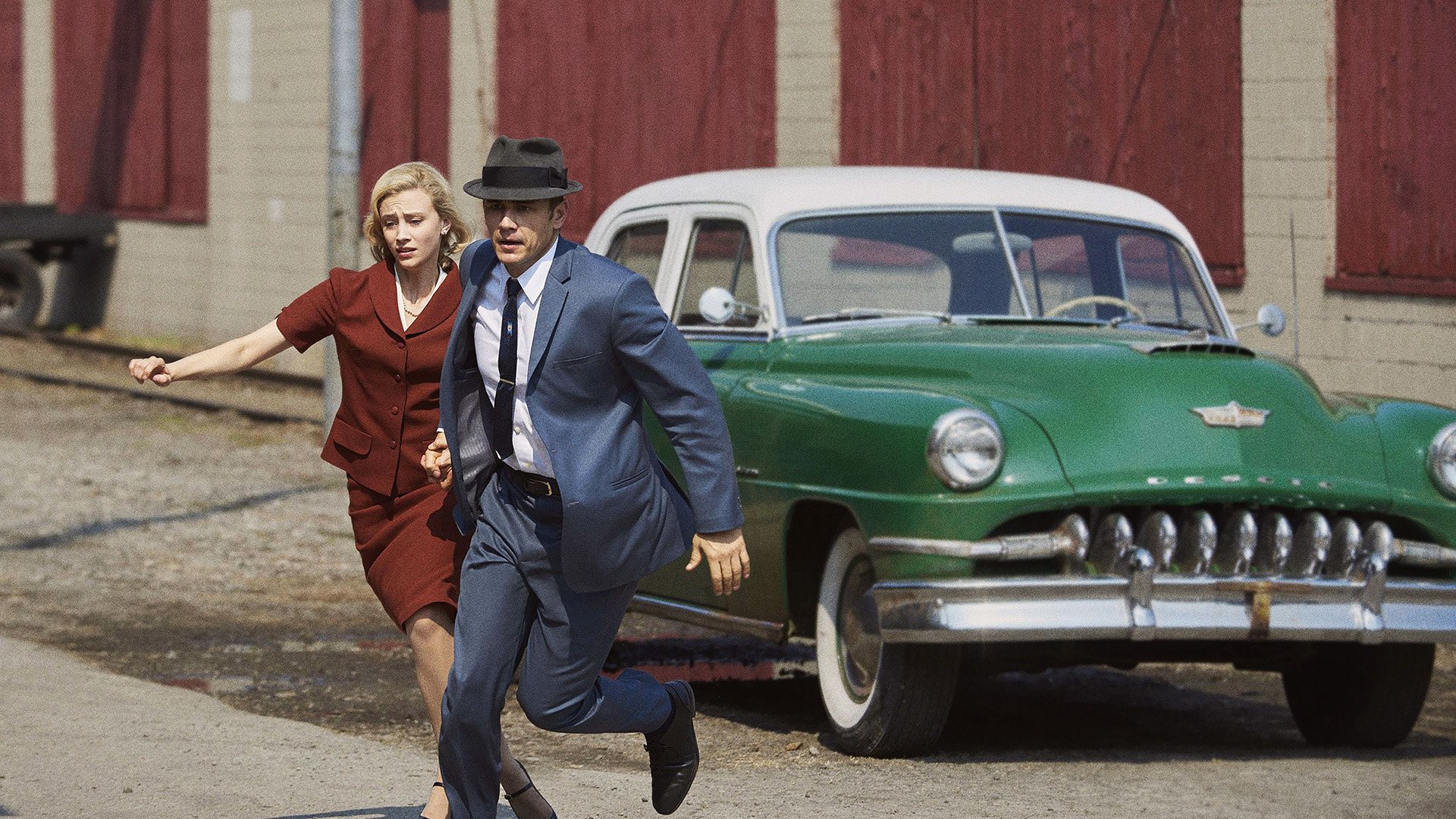 HD PC desktop wallpaper from TV show 11.22.63: a man in a suit and fedora and a woman in a red dress run past a green 1950s car beside a brick building.