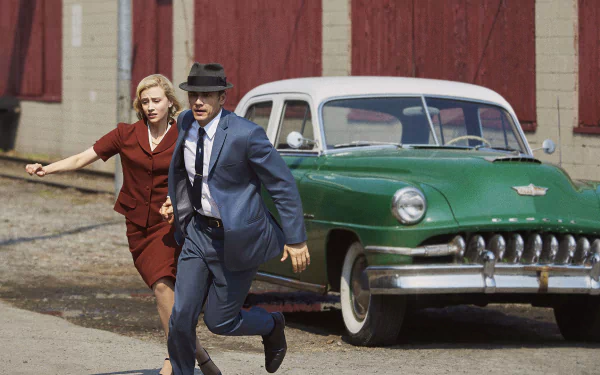 HD PC desktop wallpaper from TV show 11.22.63: a man in a suit and fedora and a woman in a red dress run past a green 1950s car beside a brick building.