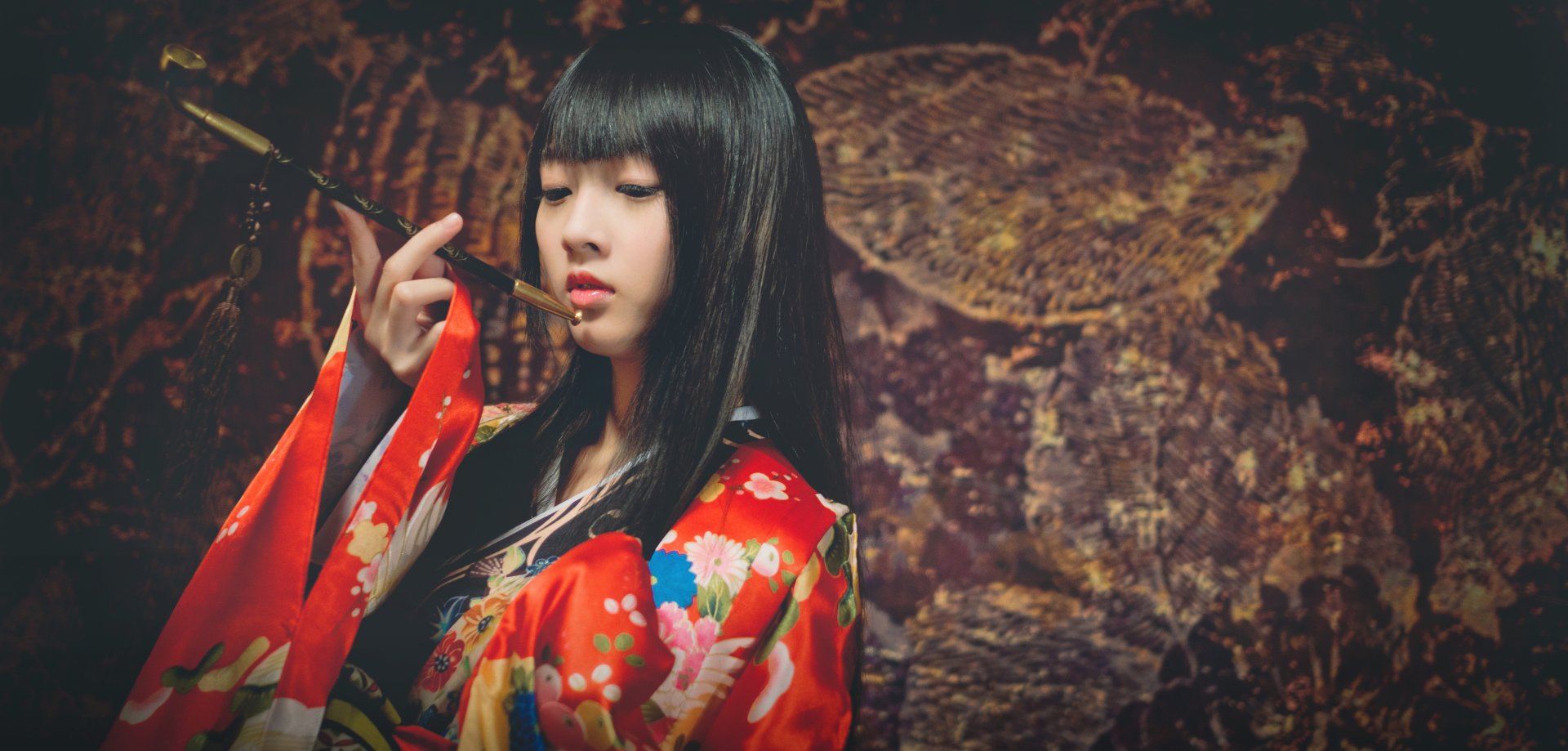 A brunette Asian woman gracefully poses in a vibrant kimono, set against a richly textured background. This striking image captures elegance and cultural beauty in 4K Ultra HD.