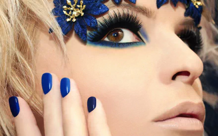 Close-up HD desktop wallpaper of a woman's face featuring striking brown eyes, bold blue makeup, matching blue nails, and decorative blue floral accents.