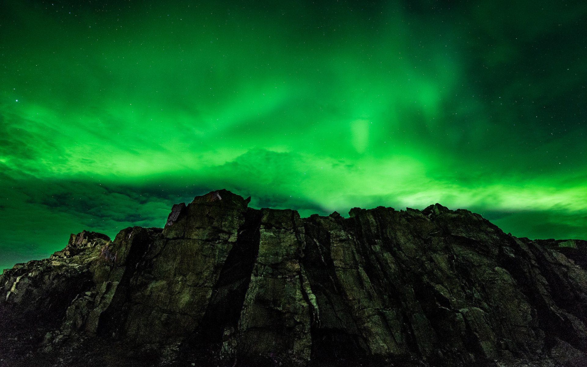 HD desktop wallpaper showing a rugged mountain silhouette beneath a vibrant green aurora borealis glowing in the night sky over natural wilderness.