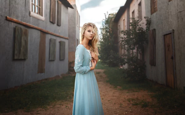 Blonde model Alice Tarasenko in a flowing blue dress stands in a narrow village street with a soft depth of field, captured in this HD PC desktop wallpaper.
