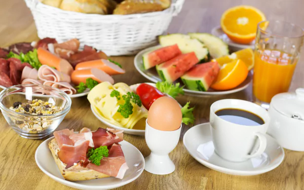 A vibrant breakfast spread with muesli, boiled egg, coffee, juice, assorted cheeses, meats, fresh fruit, and bread displayed in a bright, high-resolution still life.