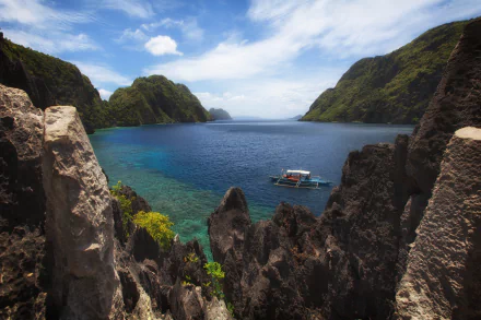 A boat floats on the clear blue ocean surrounded by towering mountains and rocky formations in a scenic Philippine coastal landscape.