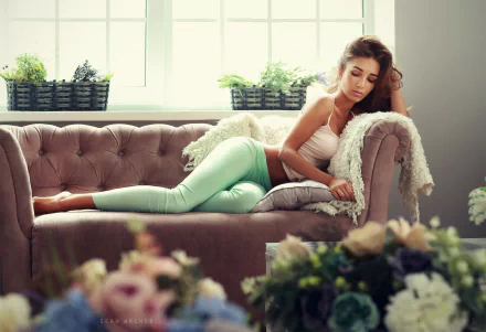 Brunette woman in jeans reclining on a tufted sofa near a bright window, surrounded by flowers, captured in an HD PC desktop wallpaper and background.