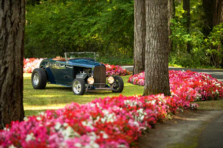 HD desktop wallpaper featuring a classic Ford Deuce Roadster hot rod parked on grass beside vibrant pink flowers and tall trees in a lush, green garden setting.