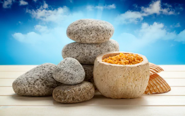 Man-made spa scene: stacked smooth stones, a bowl of orange bath salts and seashells on wooden slats against blue sky — 5K Ultra HD PC desktop wallpaper/background.