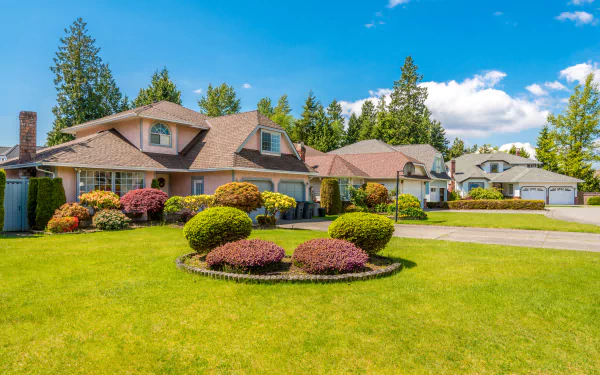 A 4K Ultra HD desktop wallpaper showing a suburban neighborhood with well-kept grass, shaped bushes, and man-made houses under a bright blue sky.