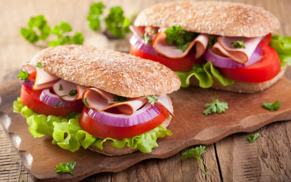A close-up of two fresh vegetable and deli sandwiches on whole grain buns, set on a wooden board. 4K Ultra HD PC desktop wallpaper featuring vibrant food.