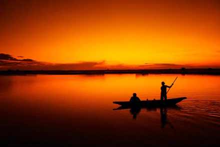 A fisherman navigates a boat through a serene lagoon in Vietnam at sunset, surrounded by vibrant orange hues reflecting on calm waters. An evocative HD wallpaper capturing tranquil beauty.