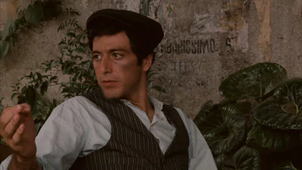 HD PC desktop wallpaper: still from the movie The Godfather — young man in period vest and cap leaning against a wall, pensive expression, leafy background
