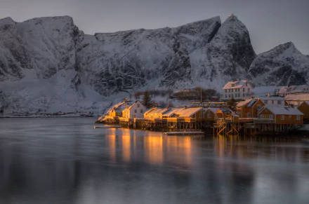 A stunning HD desktop wallpaper of Lofoten featuring snow-capped mountains and a serene coastal village bathed in warm light, reflecting on the calm waters.