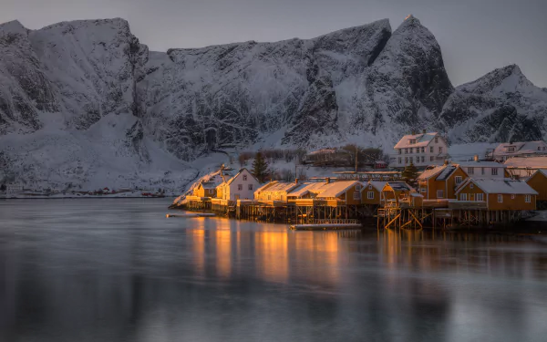 A stunning HD desktop wallpaper of Lofoten featuring snow-capped mountains and a serene coastal village bathed in warm light, reflecting on the calm waters.