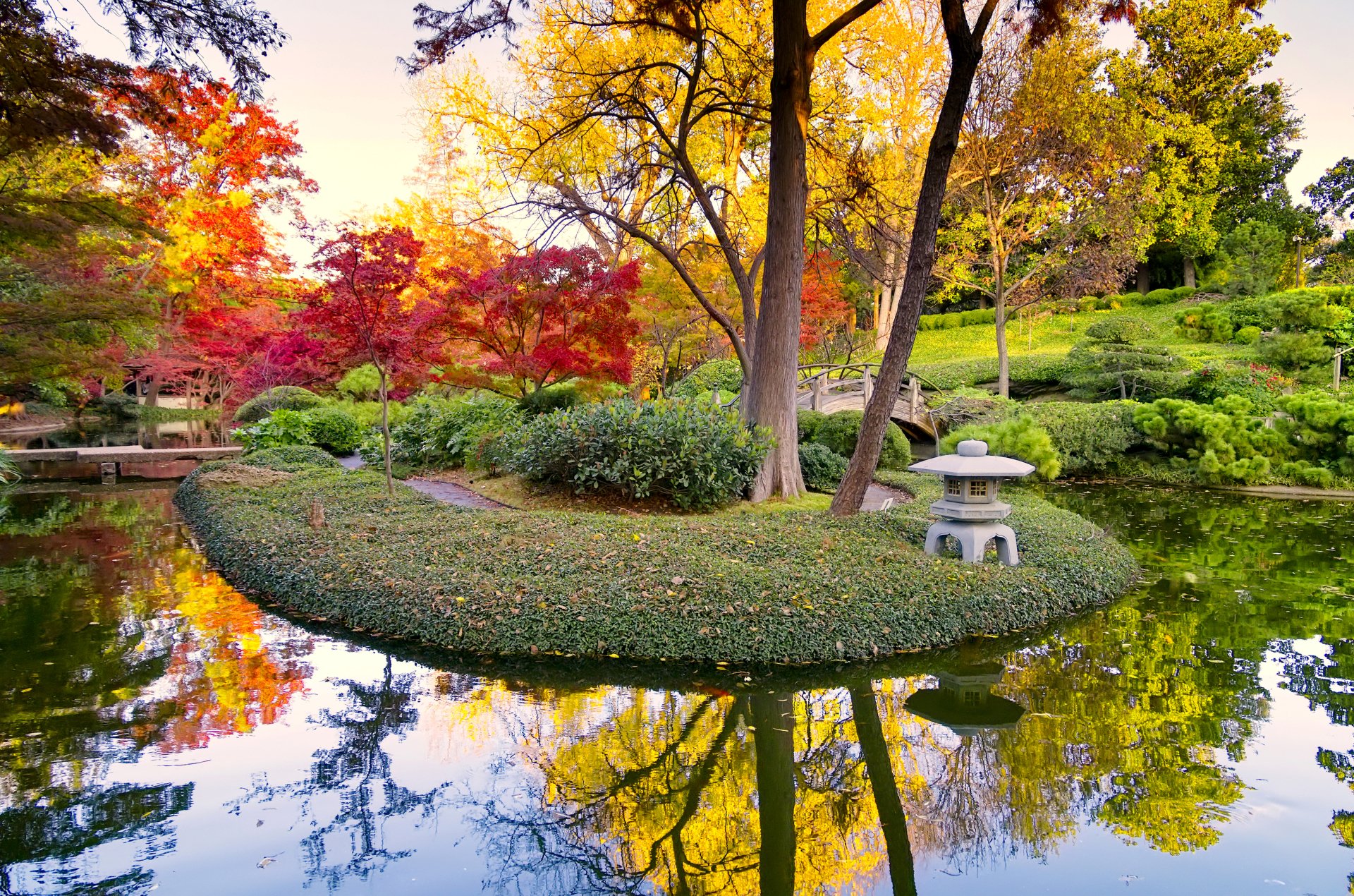 Vibrant autumn foliage reflects in a serene pond, surrounded by lush greenery and a charming stone lantern, creating a calming nature scene in the park. 4K Ultra HD wallpaper.