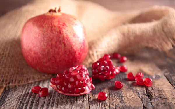 A vibrant pomegranate with scattered seeds rests on a rustic wooden surface, captured in stunning 4K Ultra HD for a vivid PC desktop wallpaper background.
