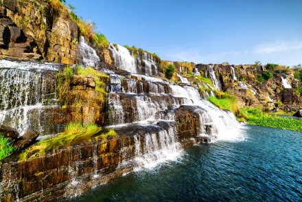 HD desktop wallpaper of a stunning waterfall in Vietnam, cascading over rocky terrain into a clear blue pool, surrounded by lush greenery under a bright blue sky.