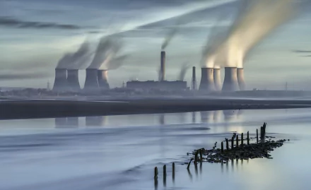 HD desktop background of a nuclear power plant emitting smoke, with its reflection visible in calm waters under a cloudy sky.