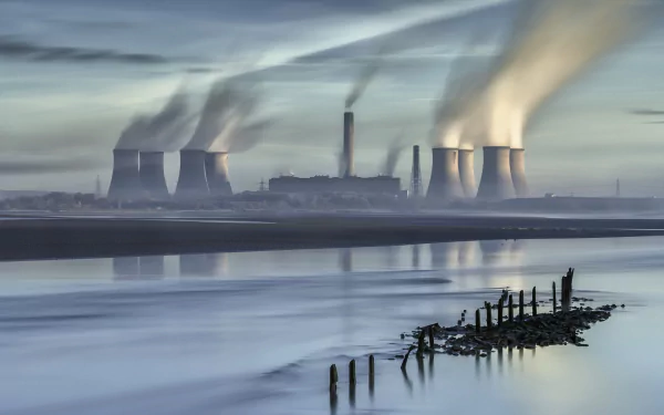 HD desktop background of a nuclear power plant emitting smoke, with its reflection visible in calm waters under a cloudy sky.