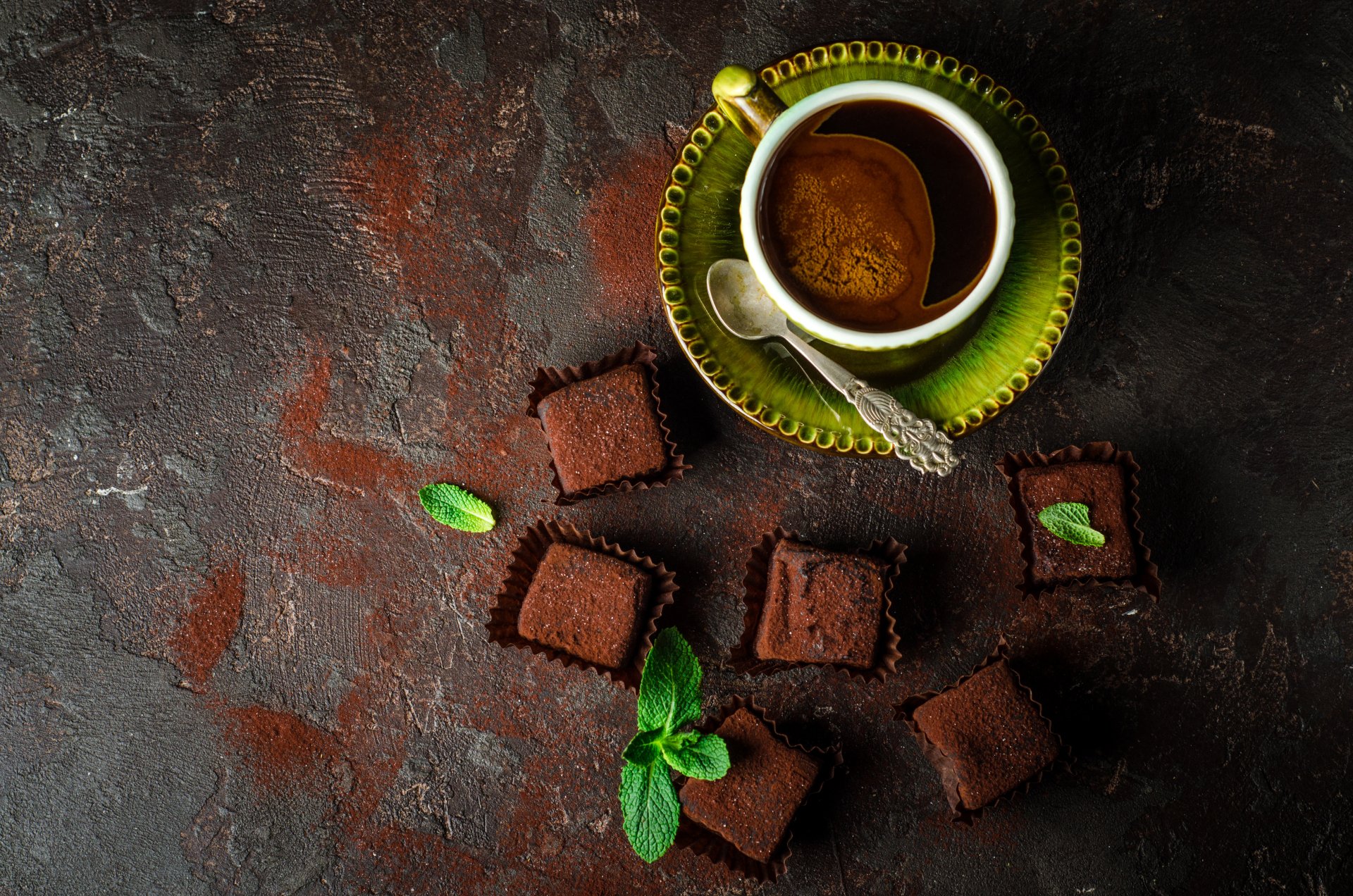 HD desktop wallpaper of a cup of coffee and chocolate sweets on a rustic background. The cup is on a green plate with a spoon, surrounded by chocolate pieces garnished with mint leaves.