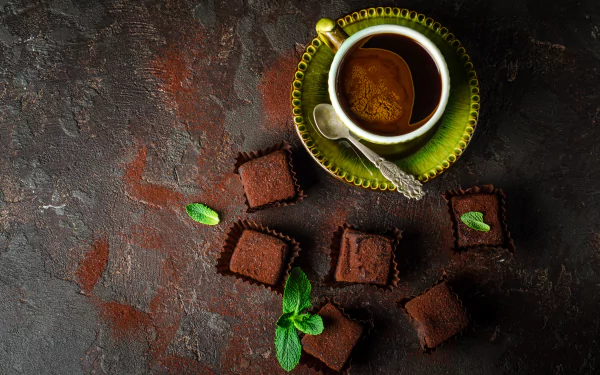 HD desktop wallpaper of a cup of coffee and chocolate sweets on a rustic background. The cup is on a green plate with a spoon, surrounded by chocolate pieces garnished with mint leaves.