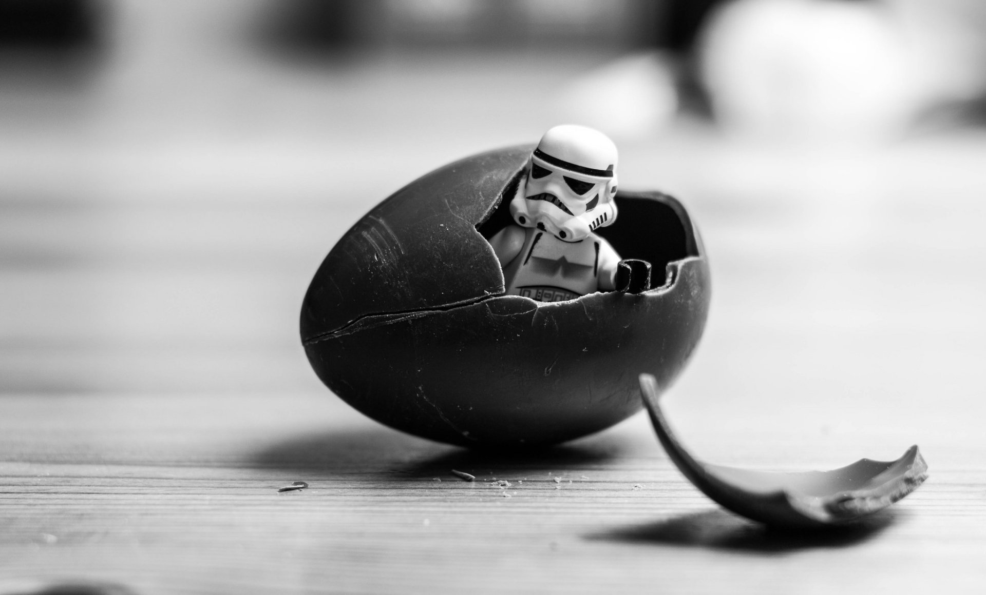 Black and white HD desktop wallpaper featuring a Lego Star Wars Stormtrooper emerging from a cracked chocolate egg on a wooden surface.
