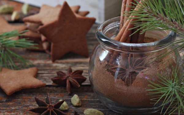 Still life HD desktop wallpaper featuring star anise, cinnamon sticks in a glass jar, spices, and star-shaped cookies with pine branches on a rustic wooden surface.