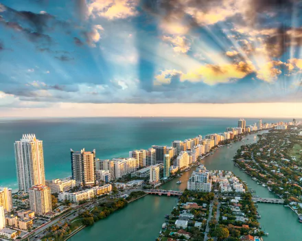 HD desktop wallpaper of Miami, Florida, showcasing a stunning cityscape along the coastline with the ocean and sunbeams breaking through the clouds on the horizon.