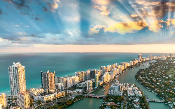 HD desktop wallpaper of Miami, Florida, showcasing a stunning cityscape along the coastline with the ocean and sunbeams breaking through the clouds on the horizon.