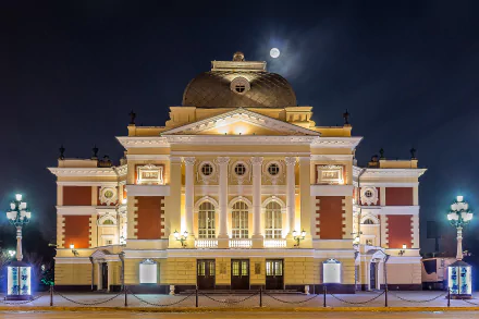 Illuminated columns of the Irkutsk Drama Theatre stand against a night sky, with the moon glowing above, showcasing stunning architecture in Russia.