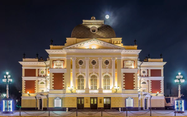 Illuminated columns of the Irkutsk Drama Theatre stand against a night sky, with the moon glowing above, showcasing stunning architecture in Russia.