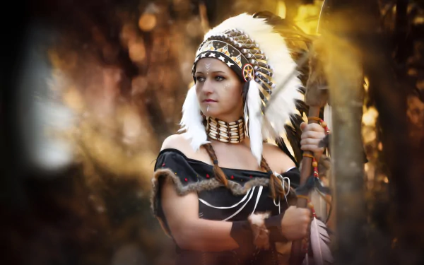 A woman in a Native American-inspired costume and headdress stands holding a spear, focused and deep in thought. The blurred background and soft feathers add to the dramatic mood of this HD desktop wallpaper.