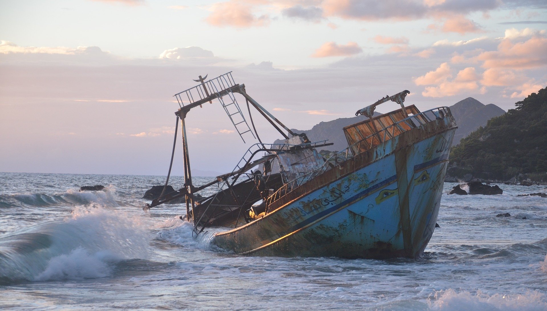 Rusting shipwreck rests on the rocky coast of Greece as ocean waves crash around it under a soft pastel sky, captured in a high-definition desktop wallpaper.