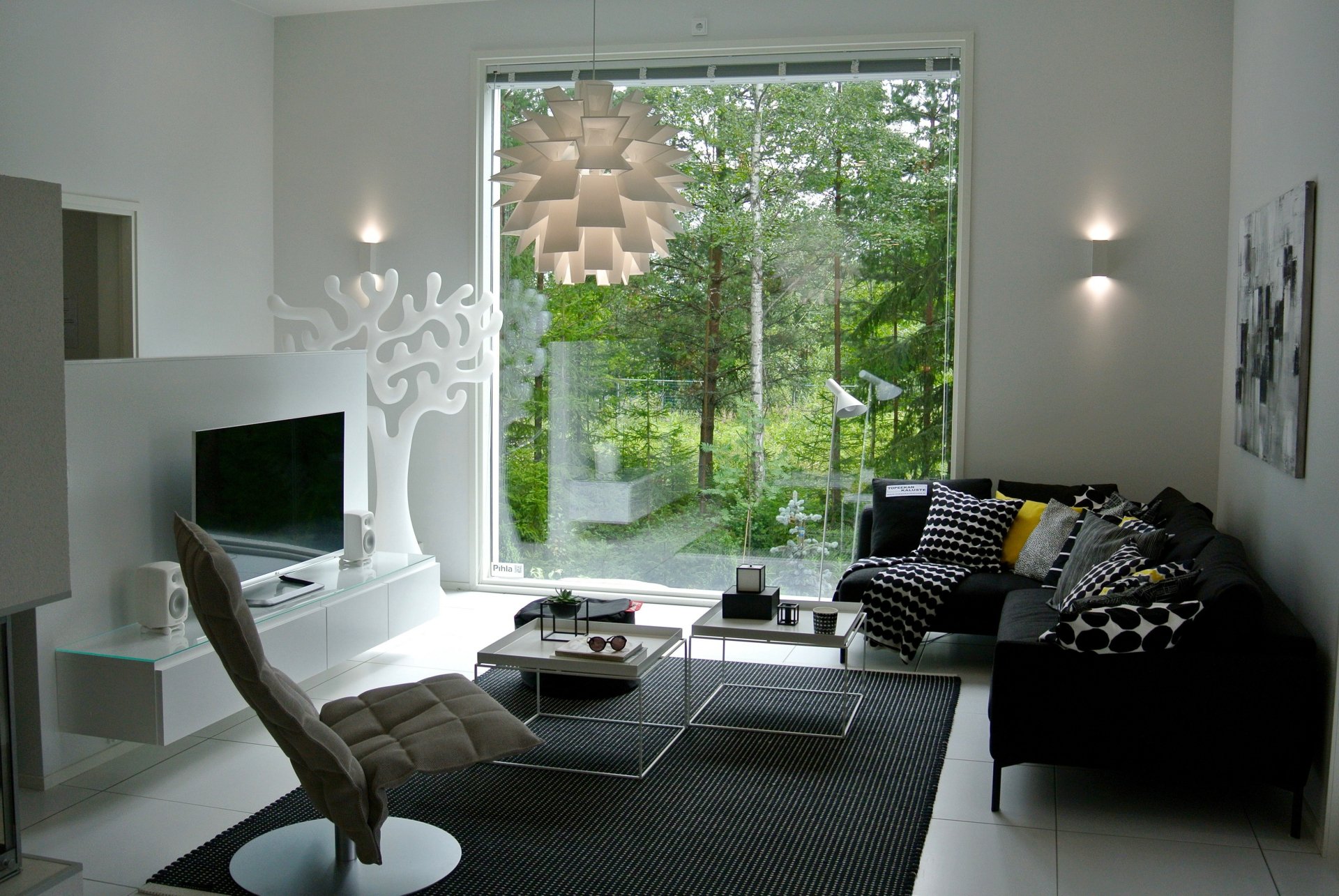 Modern living room with stylish furniture, featuring a TV, cushioned chair, and decorative cushions. Large window offers a serene view of greenery, enhancing the room's relaxing ambiance. HD wallpaper.