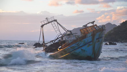 Rusting shipwreck rests on the rocky coast of Greece as ocean waves crash around it under a soft pastel sky, captured in a high-definition desktop wallpaper.