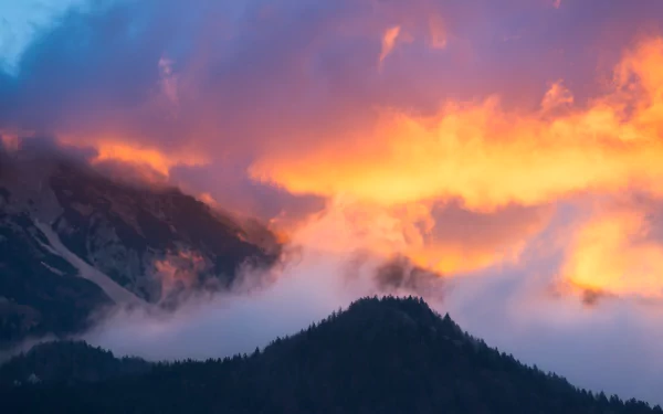 HD desktop wallpaper capturing a serene Slovenian mountain at sunrise, with fog rolling over dark peaks beneath a vibrant sky of clouds and warm morning light.