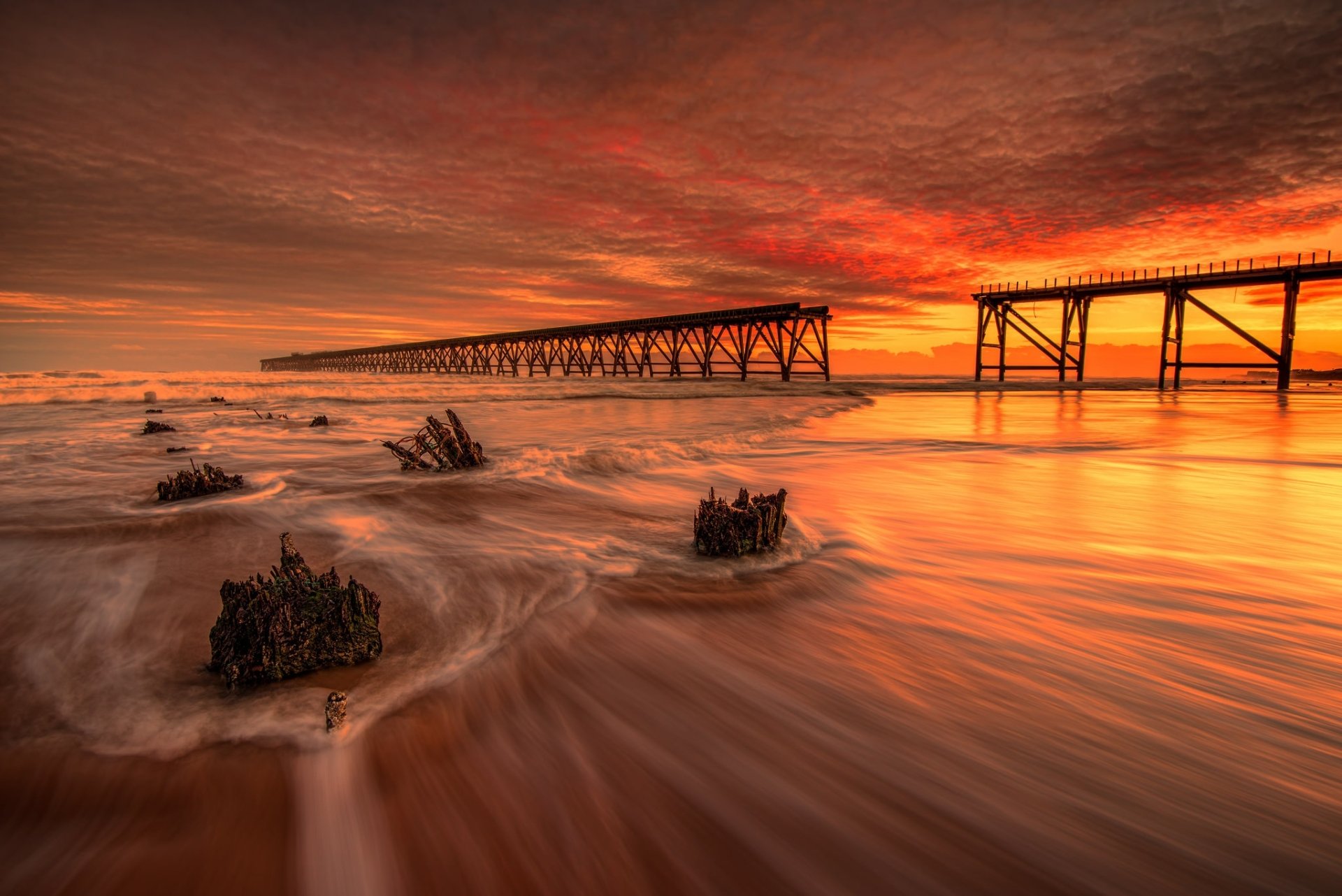 A vibrant orange sunset sky over the ocean with foam along the shore and a long wooden pier stretching toward the horizon in this HD desktop wallpaper.
