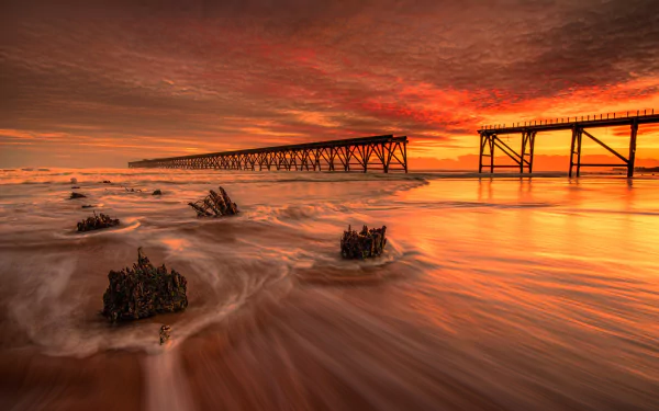 A vibrant orange sunset sky over the ocean with foam along the shore and a long wooden pier stretching toward the horizon in this HD desktop wallpaper.