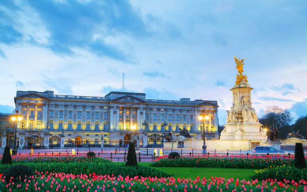 4K Ultra HD view of Buckingham Palace in London with illuminated facade, Victoria Memorial statue, and blooming tulip flowers in the foreground under a blue sky.