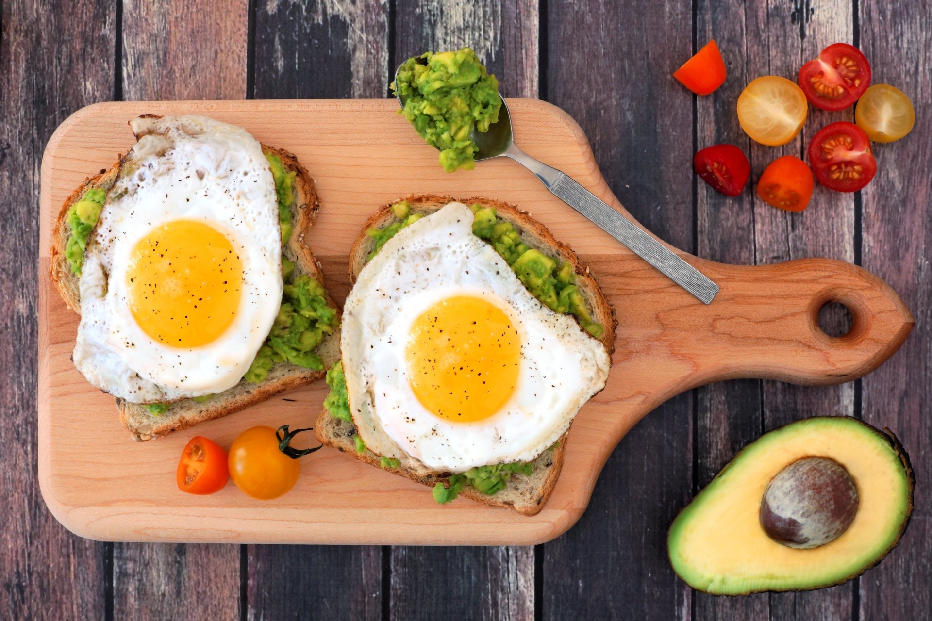 5K Ultra HD PC desktop wallpaper still-life food scene featuring avocado toast topped with sunny-side-up eggs on a wooden board, cherry tomatoes and a halved avocado.