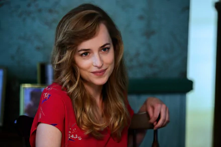 HD desktop wallpaper of Dakota Johnson smiling in a relaxed setting, wearing a red top, with a blue wall and framed photos in the background.