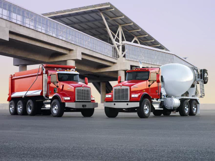 HD PC desktop wallpaper: two red Kenworth heavy trucks, a dump truck and a cement mixer, parked beneath a modern overpass under a pale sky.