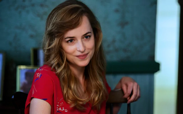 HD desktop wallpaper of Dakota Johnson smiling in a relaxed setting, wearing a red top, with a blue wall and framed photos in the background.