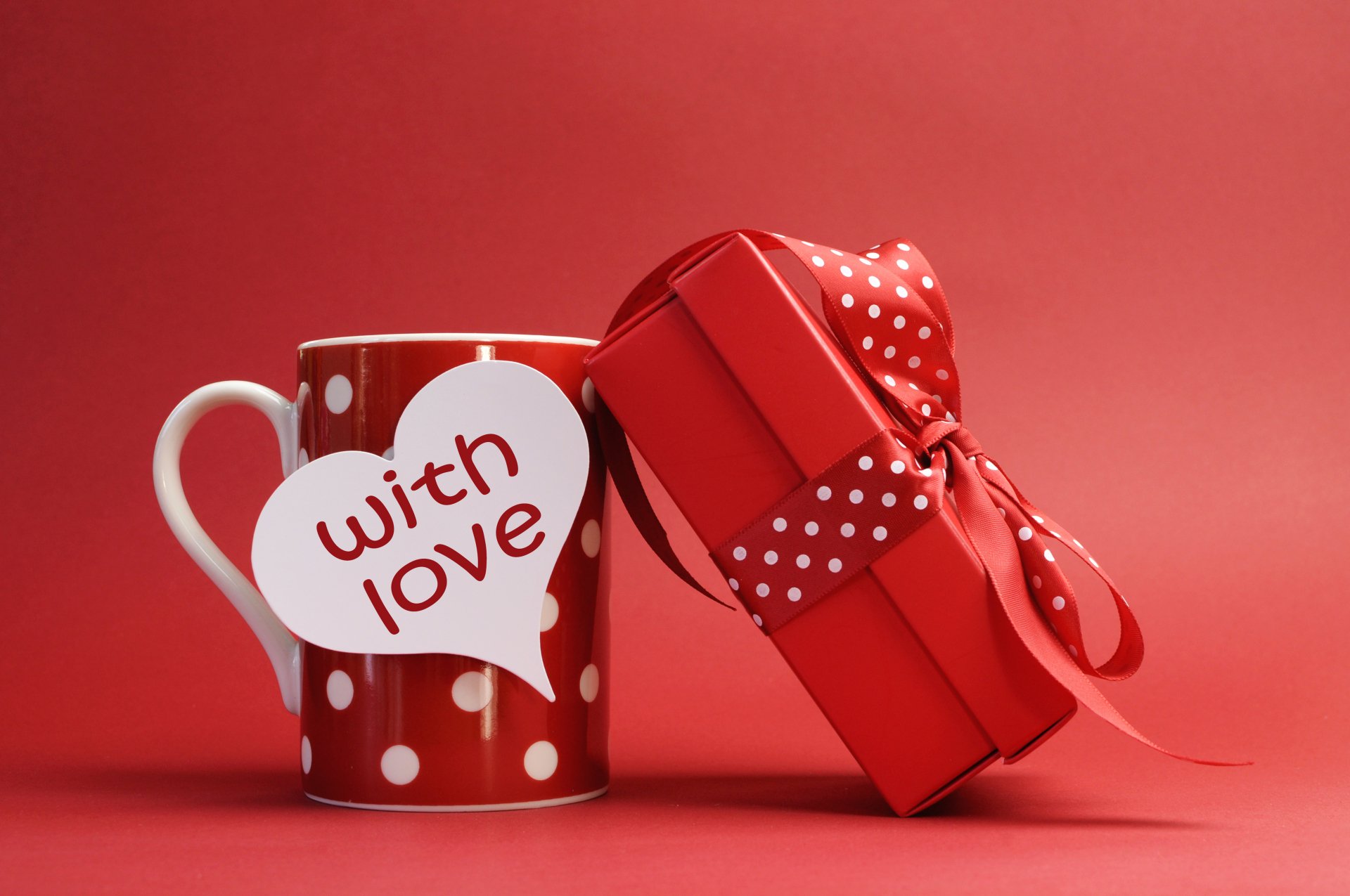 A red polka-dot cup with a heart-shaped tag saying with love next to a red gift box with a ribbon, set against a red background, themed for Valentine's Day.