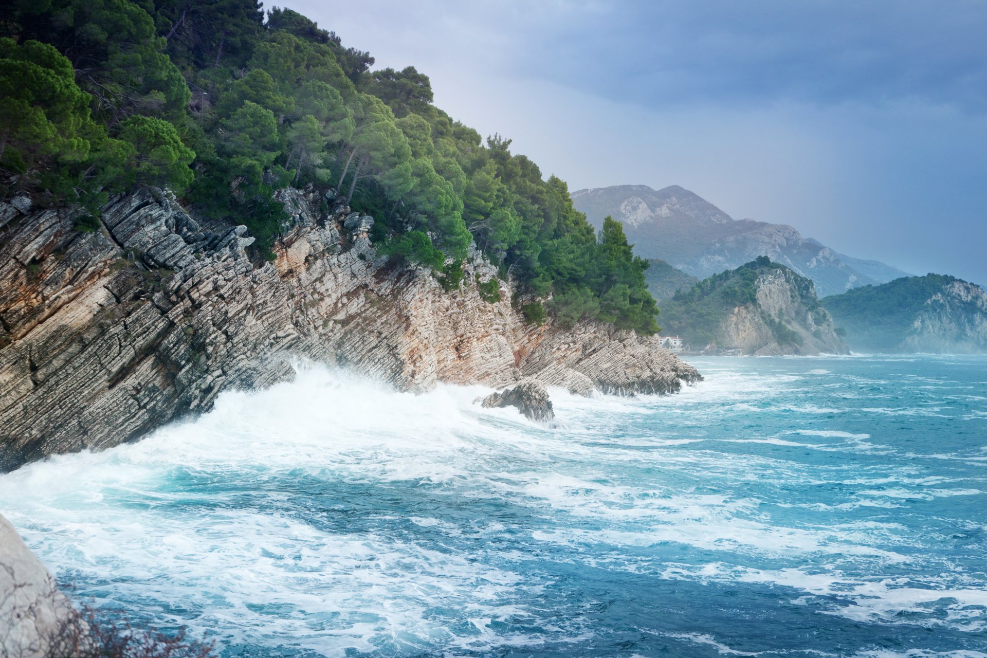 4K Ultra HD wallpaper of sea foam crashing against rugged cliffs along a lush, green coastline under a cloudy sky.