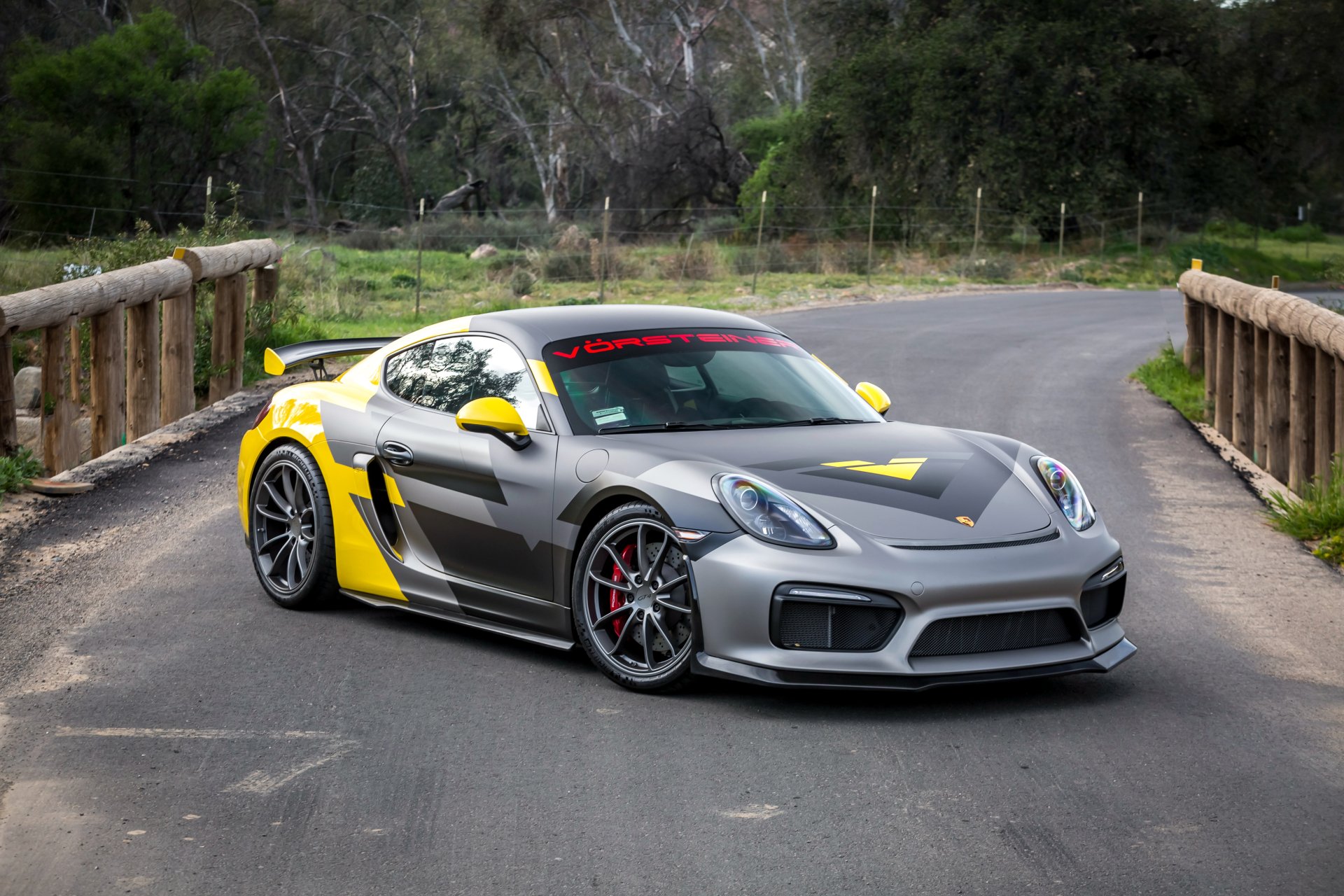 HD desktop wallpaper featuring a silver Porsche Cayman GT4, accented with yellow, parked on a rustic bridge with a scenic background.