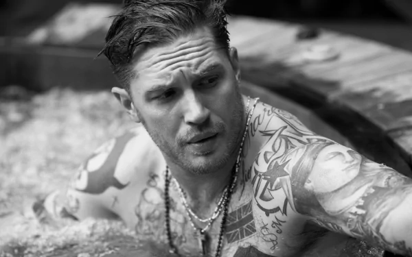 Black-and-white HD desktop wallpaper: shirtless, heavily tattooed English actor-style man emerging from water, wearing necklaces, intense gaze and moody expression.