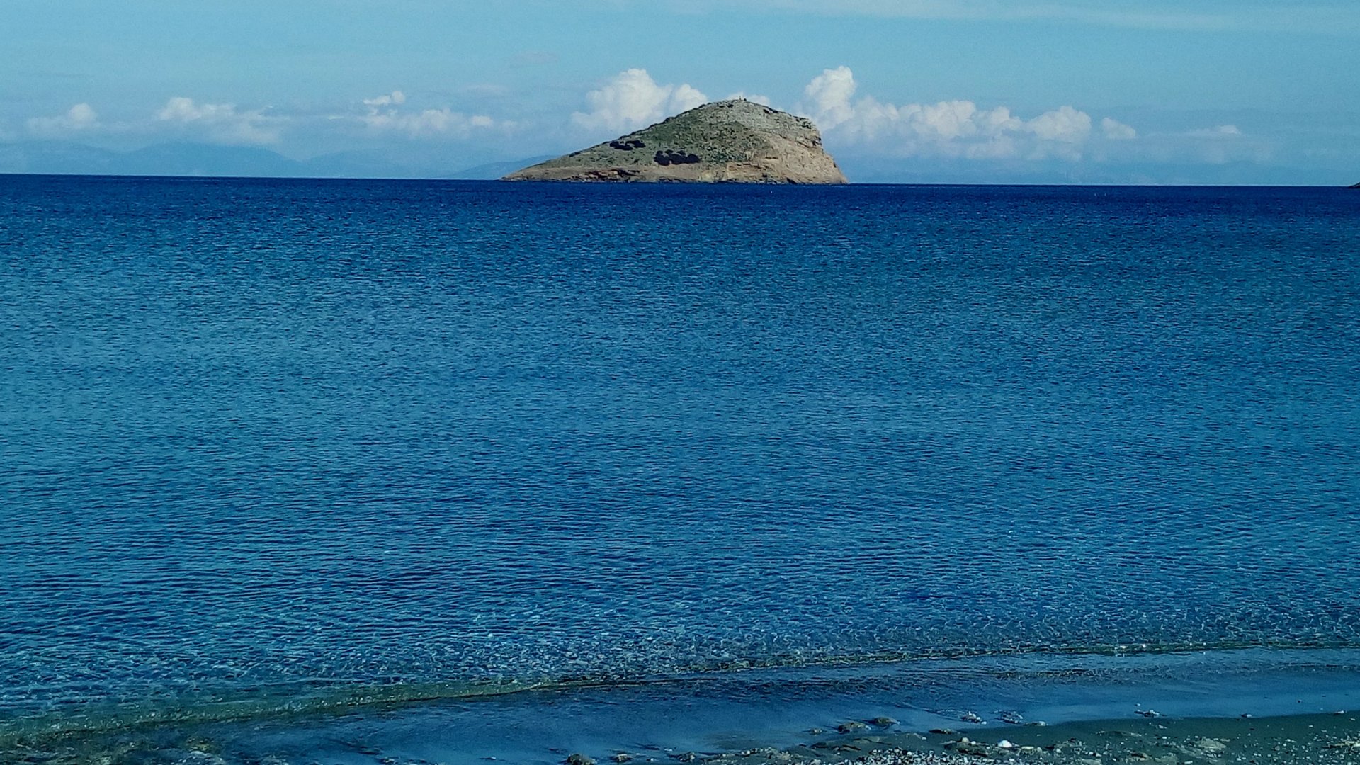 4K Ultra HD PC desktop wallpaper: tranquil Greek seashore with turquoise water and a small rocky island on the horizon beneath a clear blue sky.