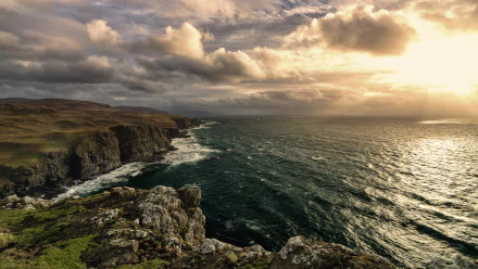 Stunning HD desktop wallpaper showcasing a dramatic coastline, with rugged cliffs meeting the ocean under a cloudy horizon and a golden sunset.