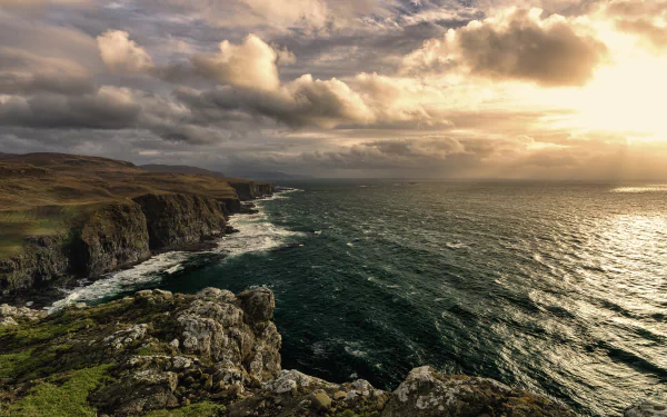 Stunning HD desktop wallpaper showcasing a dramatic coastline, with rugged cliffs meeting the ocean under a cloudy horizon and a golden sunset.