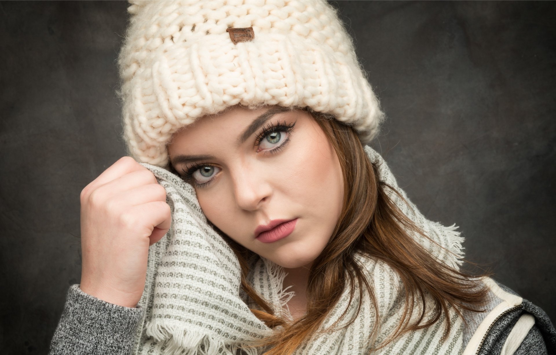 A HD desktop wallpaper featuring a brunette woman with blue eyes, wearing a cozy knit hat and a striped scarf, gazing into the camera.